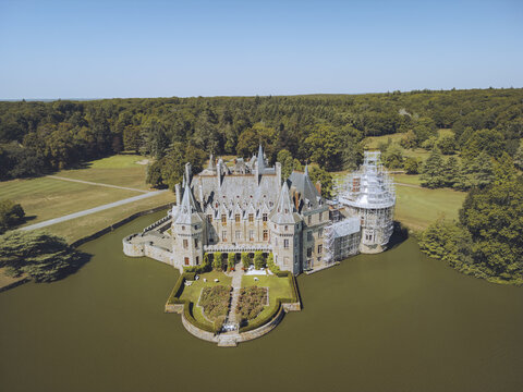 Aerial view of Ch&Atilde;&cent;teau de la Bretesche mirrored in the surrounding moat, a stone fortress amidst verdant forests, under a clear sky, Missillac, Pays de la Loire, France.