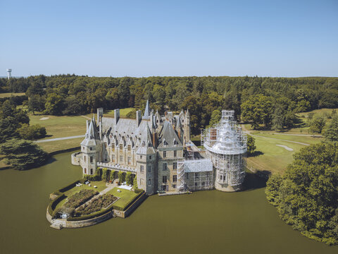 Aerial view of the Chateau de la Bretesche mirroring in the water, a tapestry of stone and nature's hues under the expansive sky, Missillac, Pays de la Loire, France.