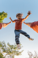 Excited young boy midair, smiling joyfully while being tossed up by a parent on a sunny day with blue sky and trees. POV view, a father high throws and catches his laughing son in a park.