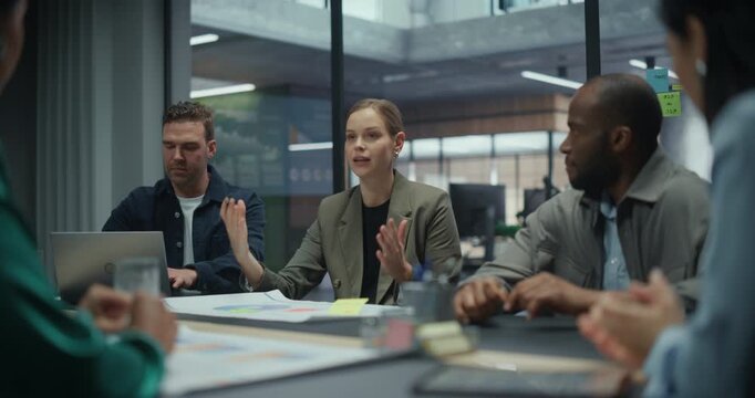 Female Executive Talking with Her Team in a Meeting, Sitting with Colleagues Behind a Table Filled with Computers and Papers. She Outlines Key Objectives while the Group Listens