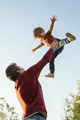 Father and son playing in the park together, dad tossing his happy laughing boy high in the air, having fun and active family time. Happy childhood or Fathers day concept. Space for text