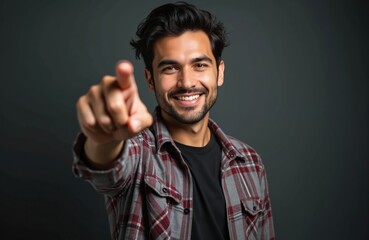 Obraz premium Attractive Hispanic man confidently points forward in studio portrait. Wears plaid shirt over black t-shirt, making eye contact with determined expression. Casual attire, dark background, bearded