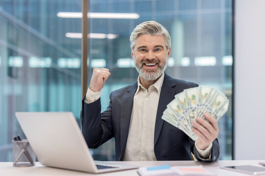 Mature businessman in suit clutching a fan of hundred-dollar bills and pumping his fist at a laptop-covered office desk. Celebrating financial success. Wealth and confident entrepreneurial achievement