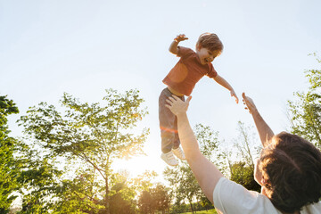 A father throws his laughing son into the air during playful family time at a park, enjoying the outdoors together at sunset. Space for text