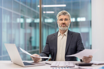 Businessman looking shocked and overwhelmed while reviewing financial papers and dealing with...