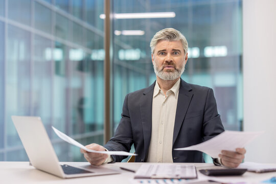 Businessman looking shocked and overwhelmed while reviewing financial papers and dealing with stress, debt, and economic problems in a modern office setup - Powered by Adobe