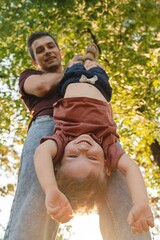 Playful dad holds his son upside down in a sunny park, as the boy laughs joyfully surrounded by green trees and sunlight. Young father having fun while playing with his son outdoors.