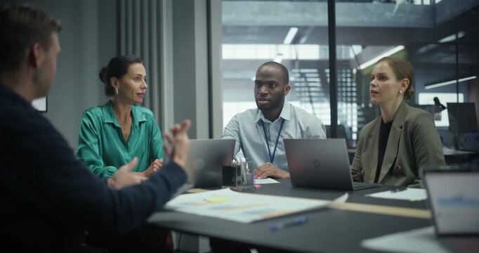 Team of Executives Talking About Business in a Meeting, Sitting Together Behind a Table Filled with Computers and Papers. Colleagues Outline Key Objectives while the Group Listens