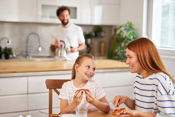 A mother and daughter share laughter while eating pizza at a kitchen table. A man stands in the background, preparing tea, creating a warm and inviting atmosphere.