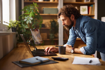 A man with curly hair and a beard is engaged in work on his laptop in a warm home office. Natural...