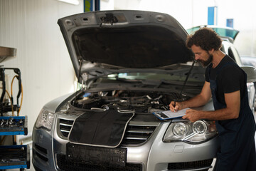 A car mechanic examines the engine of a silver vehicle while writing notes on a clipboard in a busy repair shop. Tools are visible in the background.