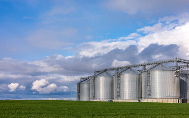 Aerial view of large silos with blue roofs, situated in a rural landscape. The silos are used for grain storage, surrounded by fields and trees under a cloudy sky.