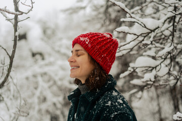 young woman smiles warmly, wearing a red hat and scarf, with snow falling around her