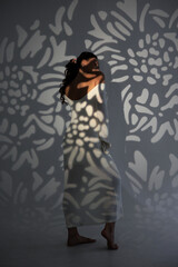 Woman in white dress surrounded by floral shadows on a wall in soft lighting