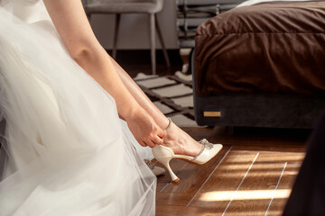 Beautiful bride wearing wedding dress and shoes sitting on sofa in room