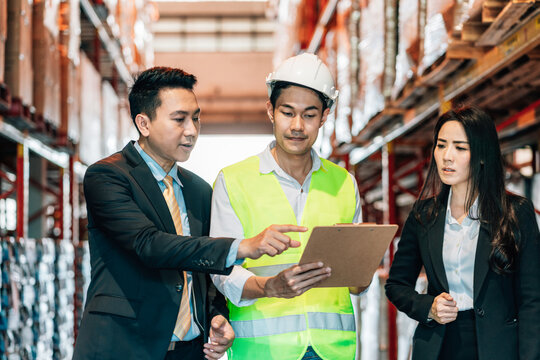 business man and woman in a suit smiles as he talks to a warehouse worker in a hard hat and safety vest, holding a clipboard. They are in a warehouse surrounded by tall shelves of merchandise