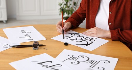 Calligraphy. Woman with brush writing phrase Keep Going on sheet of paper at wooden table indoors, closeup