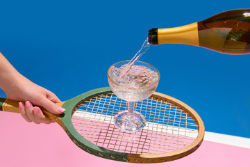 Champagne pouring into coupe glass on vintage tennis racket over pastel pink and blue background. Concept of alcohol campaigns, premium sport clubs, and event promotion visuals.