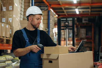 In white hard hat, scanning the box. Man in uniform is working in the storage with packages for shipment