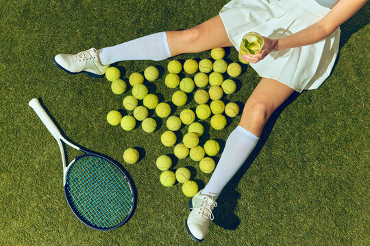 Overhead view of tennis player legs with cocktail in hand, racket and balls on grass court. Concept of premium sport clubs, alcohol advertising, and social media content creation. - Powered by Adobe