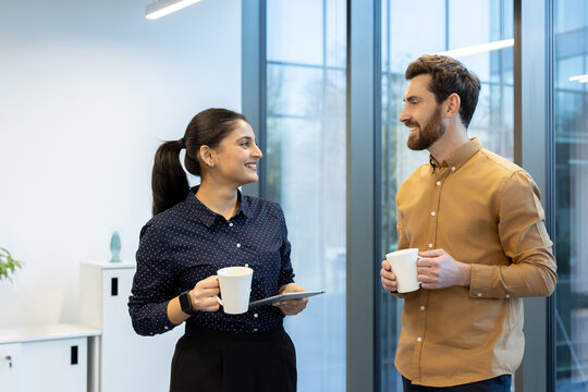 Two diverse business colleagues engaging in a friendly discussion and collaborating, holding coffee mugs during a casual break in a modern office environment - Powered by Adobe