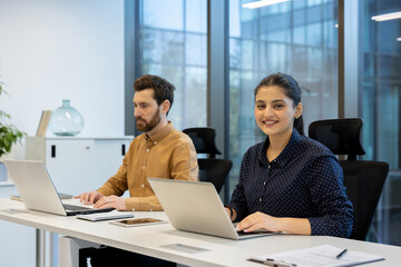 Multiracial business professionals are actively working on laptops at a shared desk in a contemporary open-plan office, highlighting teamwork and focused productivity in a modern workplace setting