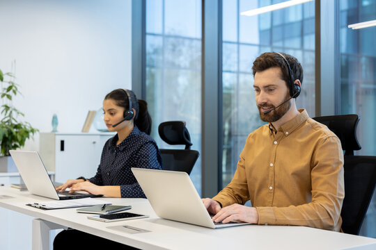 Two diverse call center operators wearing headsets and using laptops while working at desks in a bright, contemporary open-plan office, providing efficient telemarketing and online technical support