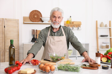 Elderly man cooking salad at white marble table in kitchen