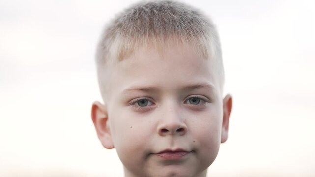 Closeup boy face looking straight with calm neutral expression and soft skin detail blond hair ear visible pale sky background portrait closeup eye focus child innocent gaze subtle smile detail