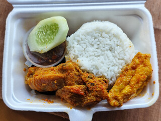 Styrofoam boxed rice with side dishes of chicken rendang, tofu, chili sauce and cucumber slices in a styrofoam box placed on a wooden table.