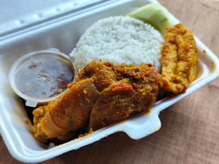 Styrofoam boxed rice with side dishes of chicken rendang, tofu, chili sauce and cucumber slices in a styrofoam box placed on a wooden table.