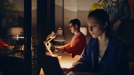 Overtime team typing laptops in lamp light coworking office. Serious colleagues
