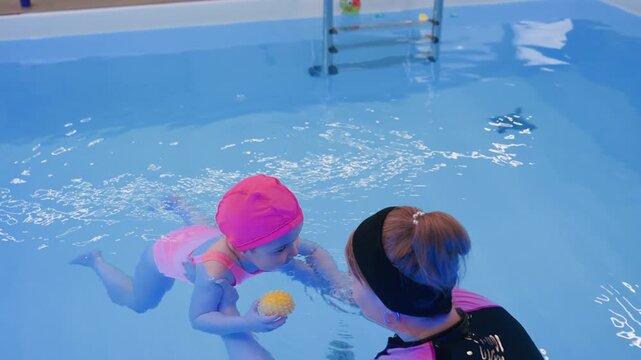 white girl in pink swim suit floats on pool water with instructor hands guiding movement supportive coaching calm toddler practice safe learning bright light reflections ripples early swimming skill