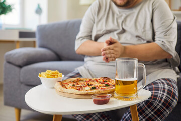 Overweight man breaks diet with pizza, beer. Fries and sauce sit on a table as he lounges on the sofa, showing obesity and a sedentary evening at home. Unhealthy habits and weight gain.