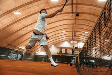 Fototapeta premium In the air, jumping. Man is playing tennis indoors in the hangar