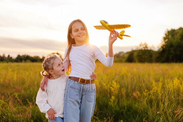 A cheerful older sister holds a toy airplane while her younger sister looks up at her with a surprised and happy expression, both standing in a sunlit field at sunset.