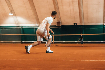 Obraz premium In white sport clothes. Man is playing tennis indoors in the hangar