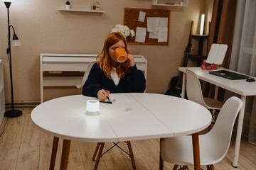 A teenage girl studies in the living room during evening hours, drinking coffee while focusing on...