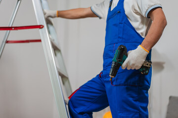 Skilled Worker in Blue Overalls Holding Drill While Standing on Ladder in Bright Indoor Setting