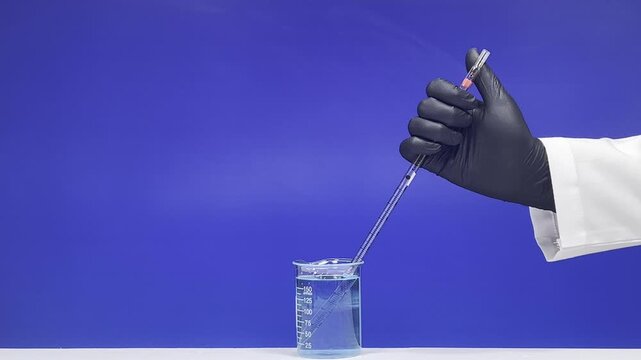 Hand pressing and emptying glass pipette into beaker with blue liquid on laboratory blue background