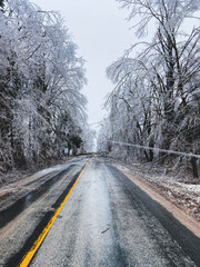 A rural road stretches into the distance, bordered by trees heavily coated in ice after a severe winter storm. A sagging power line is down across the road.