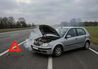 Broken down car on a rural road with engine smoke and a warning triangle for roadside assistance concept.