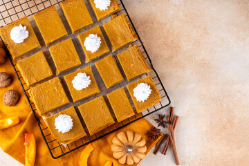 Homemade autumn taste pumpkin pie bars. Freshly baked pumpkin bars with whipped cream on it, with autumn decor and spices on beige table background, copy space 