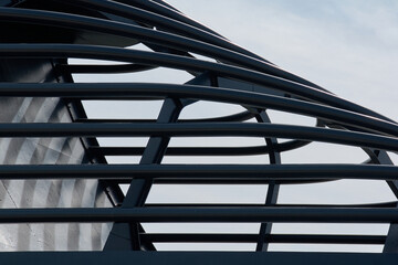 Close-up of a black metal architectural structure. The interlocking curved and straight bars create a dynamic geometric pattern against a pale blue sky.