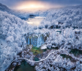 Aerial drone view of frozen waterfall and turquoise lakes surrounded by snowy forest at sunrise in Plitvice Lakes National Park, Croatia. Winter landscape with icy cascades, trail. Scenic nature