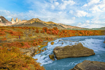 Beautiful waterfall with turquoise river and autumn foliage in Torres del Paine, Patagonia, Chile, under bright sky and mountain backdrop.