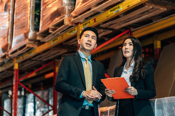 A business man in a suit gestures toward the upper shelves in a warehouse as he talks to a business woman, who holds a clipboard and looks up.