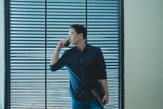 Businessman standing by window blinds making a phone call, holding a laptop, symbolizing professional communication and remote work in a modern office environment