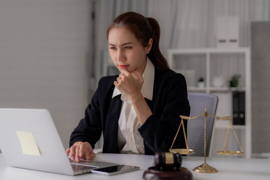 Asian adult legal professional woman concentrating analyzing documents laptop modern law office scales justice gavel thoughtful attorney lawyer paralegal