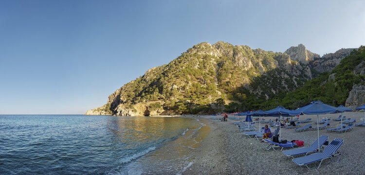 Apella beach with sun loungers and parasols by the turquoise sea against a mountain backdrop, Apella beach, Karpathos, Greece
